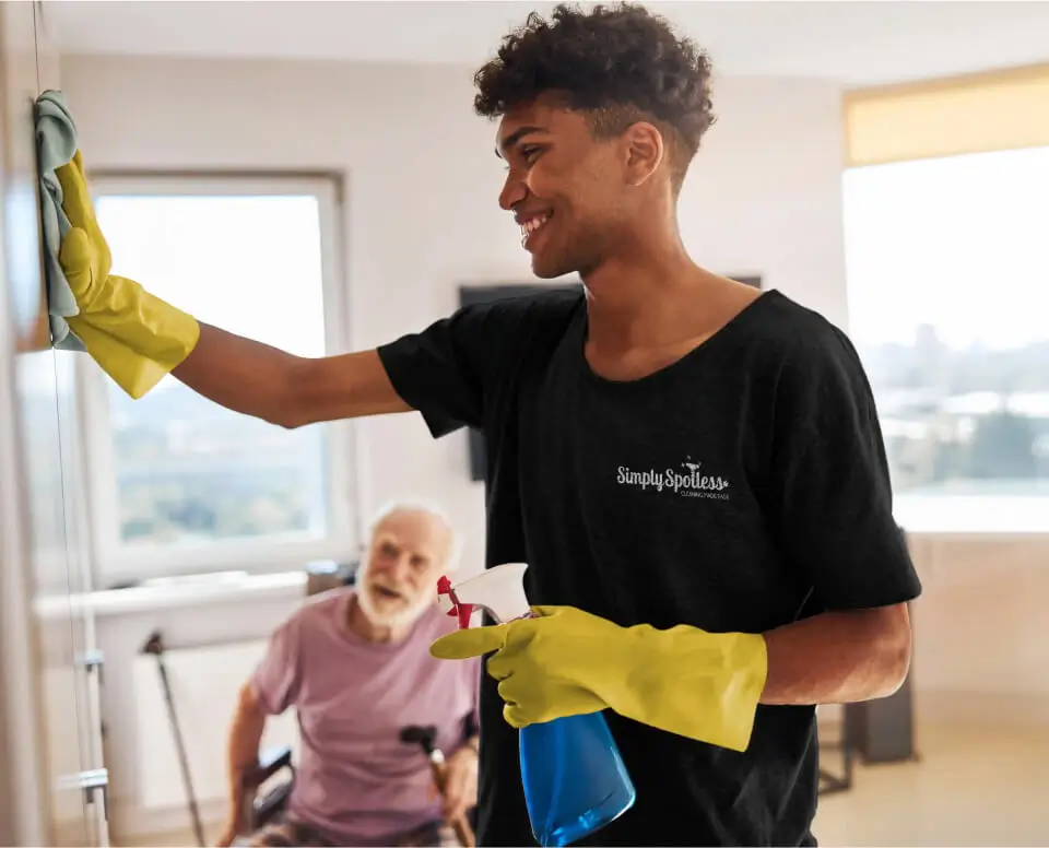 Simply spotless cleaner, cheerfully cleaning a room in an NDIS participant's home whilst the customer watches and engages